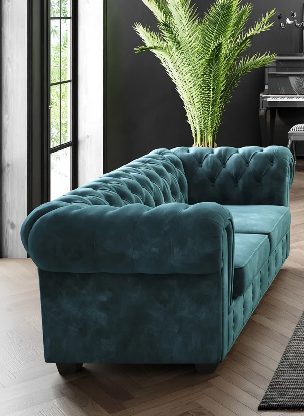 Vue latérale d'un canapé Chesterfield en velours bleu turquoise, mettant en avant son design capitonné et ses accoudoirs arrondis. Décoré avec un fond moderne comprenant une plante verte et un piano noir.