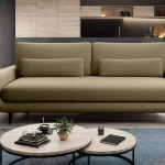 Canapé moderne en tissu olive, aux lignes arrondies, avec des coussins rectangulaires assortis, placé dans un intérieur contemporain avec une table basse ronde et un tapis bleu.