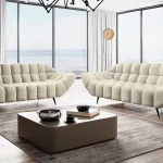 Vue intérieure d'un salon moderne avec deux canapés design beiges aux formes capitonnées et courbées, situés devant de grandes baies vitrées offrant une vue dégagée sur l'extérieur.