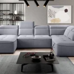 Canapé d'angle XL en tissu gris clair avec appuie-têtes réglables, dans un salon moderne avec une décoration minimaliste, étagères ouvertes, tableau abstrait et table basse noire élégante ornée de branches décoratives.