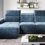 Canapé d'angle bleu moderne avec dossiers ajustables, posé sur un tapis gris, accompagné d'une table basse blanche contenant des pommes vertes. Décor minimaliste avec une étagère murale ornée de plantes et d'objets décoratifs.