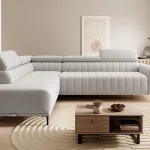 Canapé d'angle moderne en tissu gris clair avec appuie-tête réglables, disposé dans un salon lumineux avec une table basse en bois et un tapis circulaire texturé beige.