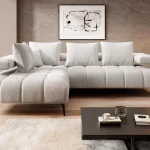 Salon moderne avec un canapé d'angle blanc texturé, décoré de coussins assortis, posé sur un parquet clair. Mur en béton ciré avec tableau abstrait, éclairage suspendu noir, lampadaire design en bois, grande baie vitrée avec vue sur la nature, et table basse foncée sur un tapis beige.